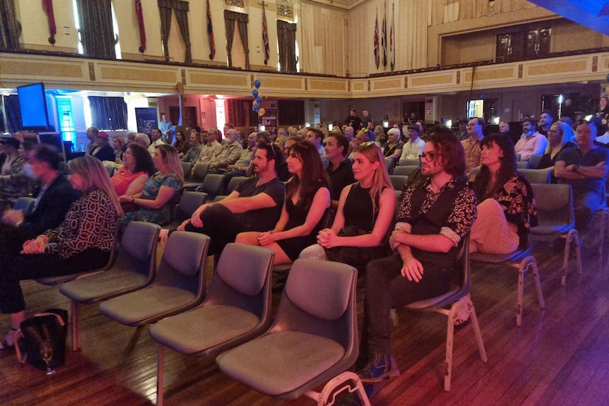 A crowd sits in seats inside a Town Hall.