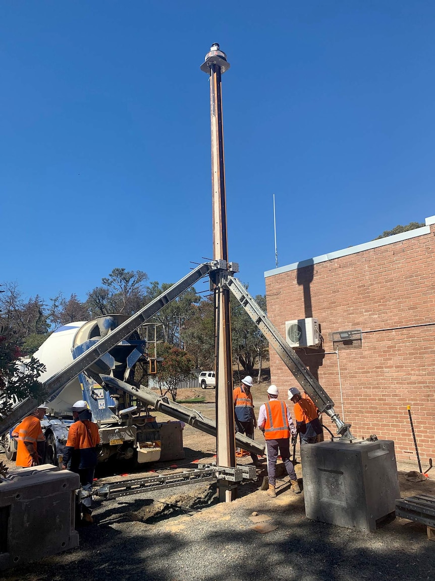 Workmen erect a stobi pole next to a brick building.