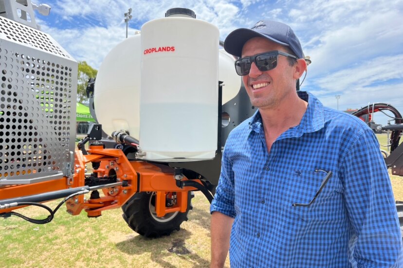 A young man wearing a hat and sunnies grins in front of a high tech spray rig with a big tank on it