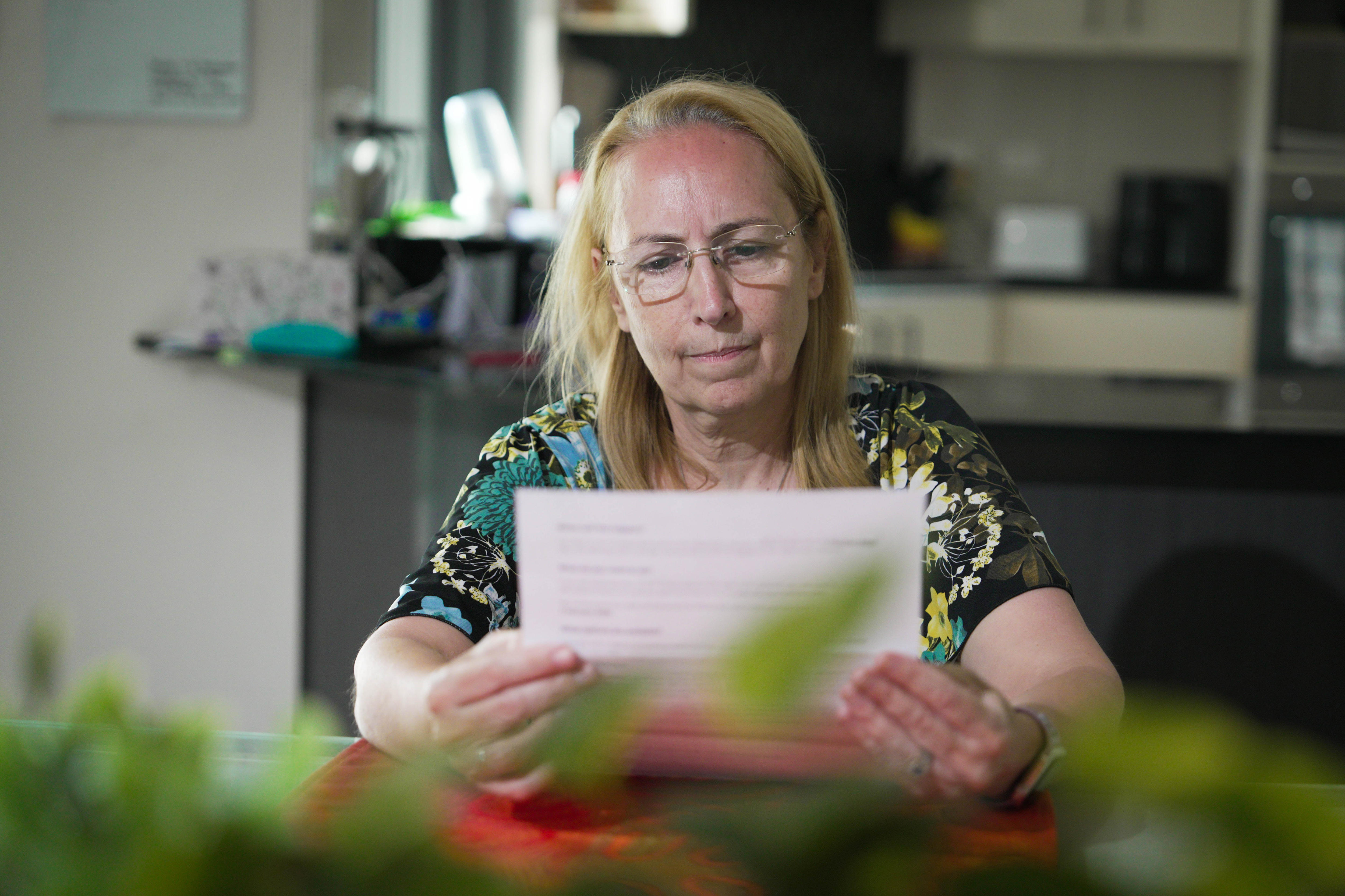 Una mujer rubia de tez blanca está sentada en la mesa de su cocina leyendo una carta, con una planta borrosa en primer plano.