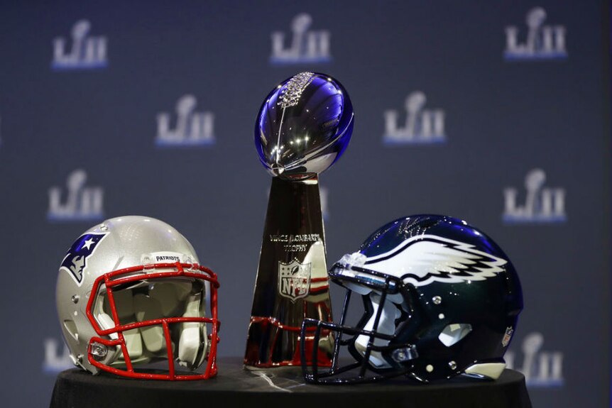 Philadelphia Eagles and New England Patriots helmets are on a table with the Vince Lombardi Trophy