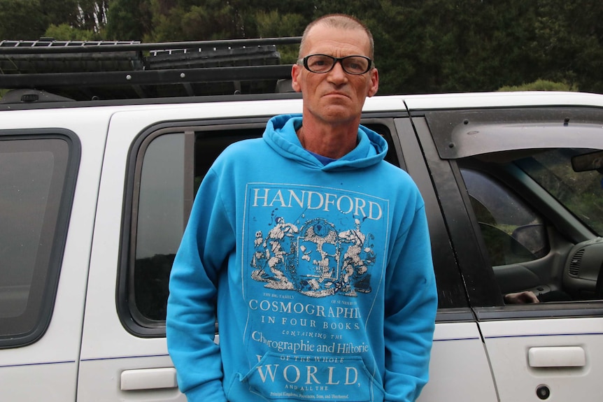 A man wearing a blue hooded jumper stands in front of his car.