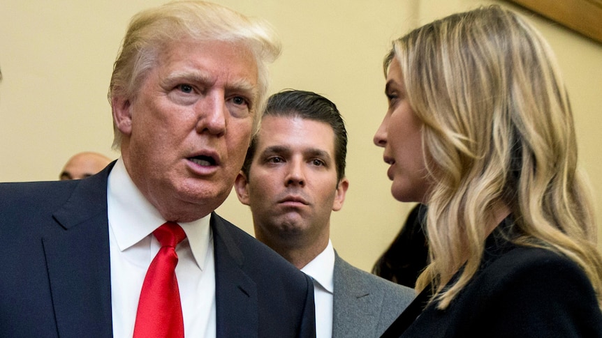 Donald Trump in a suit and red tie listens to his daughter Ivanka speak, while Don Jr stands in the background