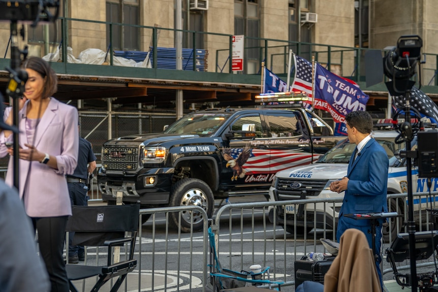 A suited reporter turns around and looks at a black truck that has pro-Trump images and slogans painted and displayed on flags