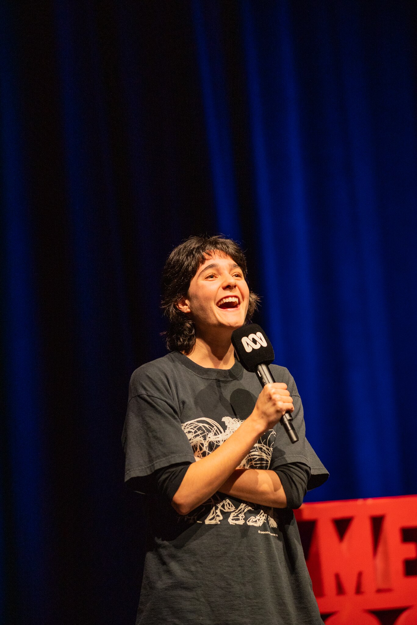 Lara Ricote on stage with a red Comedy Festival sign and dark blue stage curtains in the background.