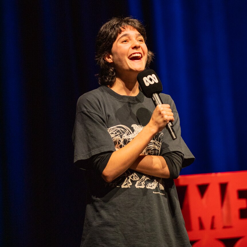Lara Ricote on stage with a red Comedy Festival sign and dark blue stage curtains in the background.