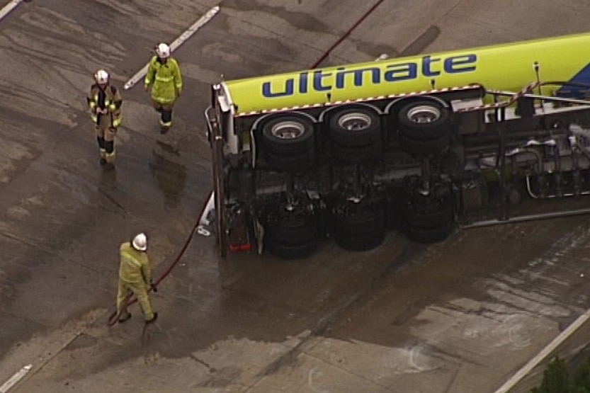 Three firefighters stand around the fuel tanker on its size on the road and one is holding a hose