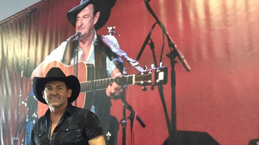 A man, wearing a black shirt and a black cowboy hat, stands in front of a large photo of Slim Dusty playing the guitar.