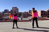 A boy and his father stand on a rooftop, each pointing forward a soccer referee flag