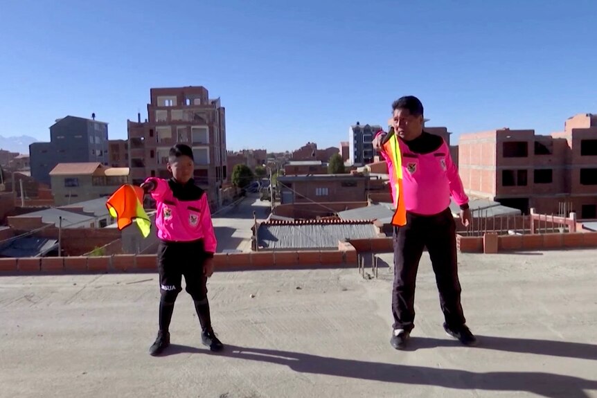 A boy and his father stand on a rooftop, each pointing forward a soccer referee flag