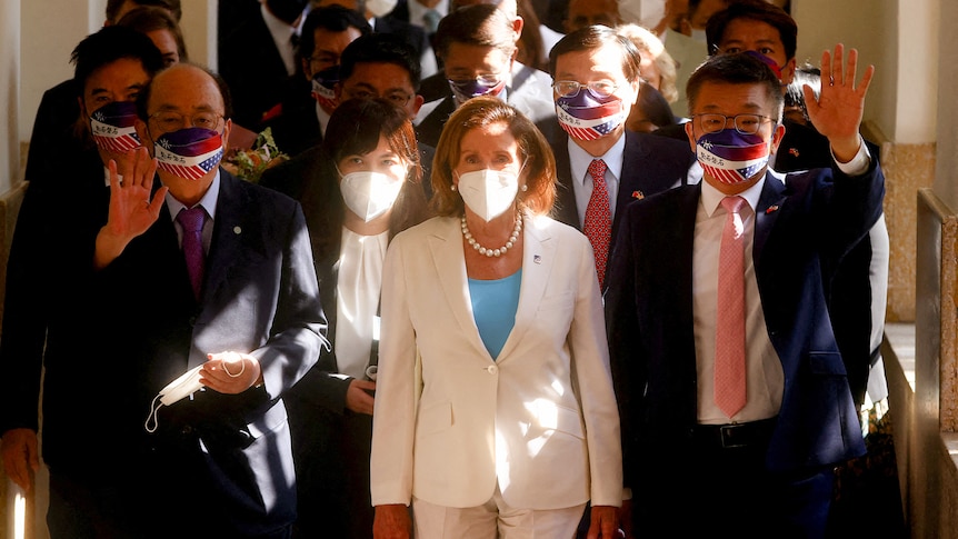 Nancy Pelosi dressed in a white pantsuit and wearing a white mask walks among a crowd of people.