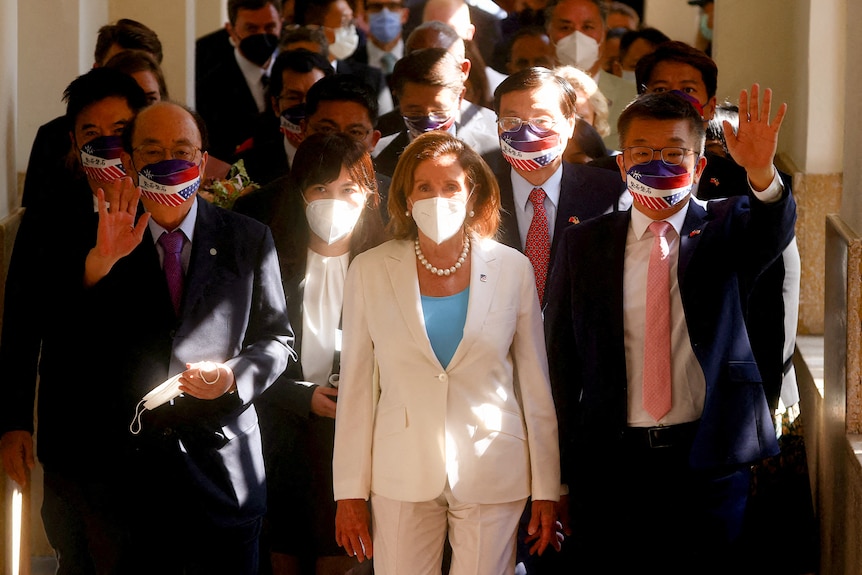 Nancy Pelosi dressed in a white pantsuit and wearing a white mask walks among a crowd of people.