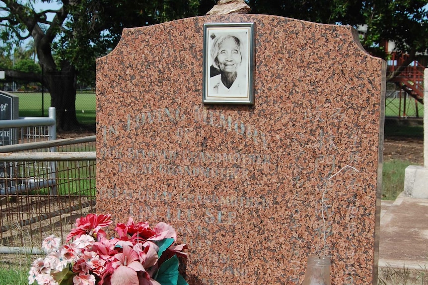 3:4 of a rose-coloured granite headstone in a cemetery. Trees in background. Flowers and vase placed