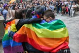 Four people put theirs arms around each other's shoulders as they stand before a crowd wrapped in a rainbow flag.