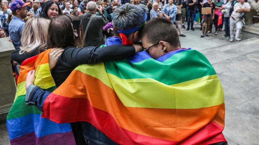 Four people put theirs arms around each other's shoulders as they stand before a crowd wrapped in a rainbow flag.