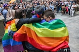 Four people put theirs arms around each other's shoulders as they stand before a crowd wrapped in a rainbow flag.