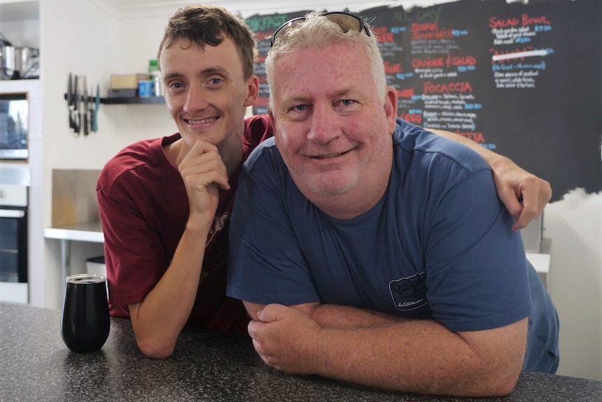 Robert, red shirt, hand under chin, smiling, arm around support worker Greg Browne, also smiling, blue shirt.