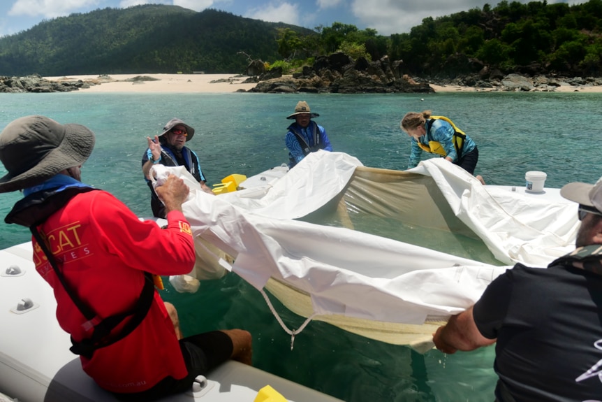 people in life jackets sit around a white square pontoon on blue water, lifting up white mech