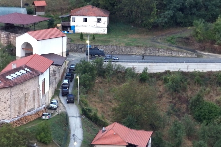 An aerial photograph shows a number of black and grey four-wheel drives and an armoured vehicle lined up on a road.