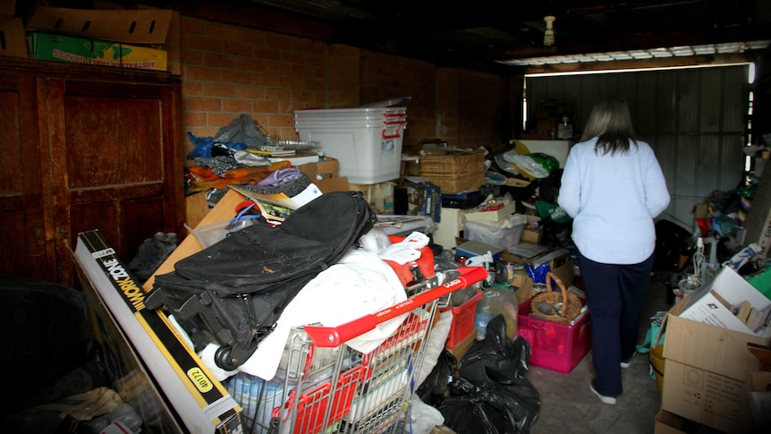 A woman stands in a garage crowded with items, including a supermarket trolley stacked with books.
