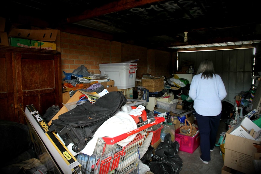 A woman stands in a garage crowded with items, including a supermarket trolley stacked with books.