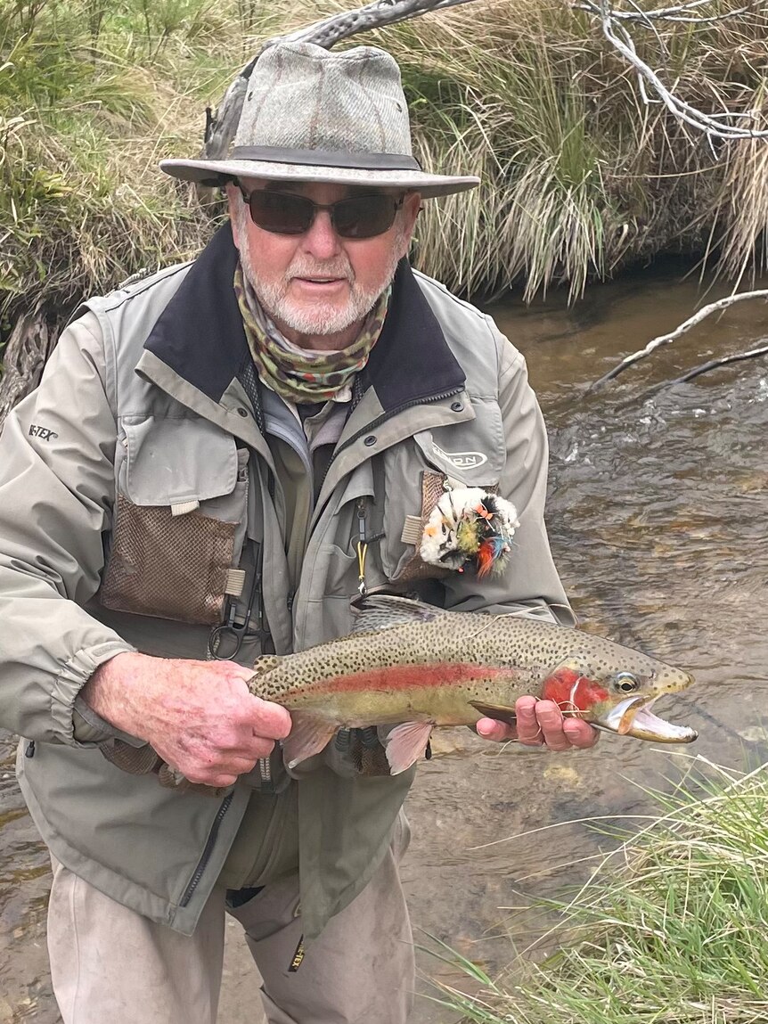 Man holding a rainbow trout fish, standing in a river. 