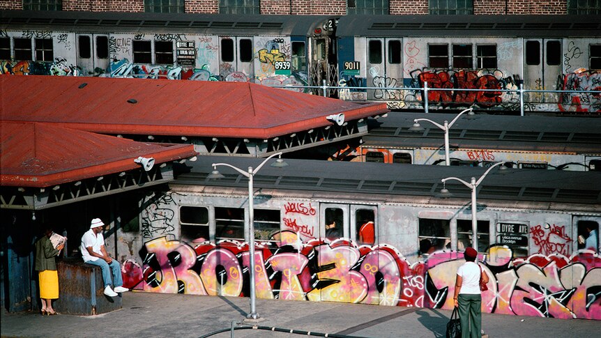 Three people wait for on subway station near three subway cars covered in colourful graffiti.