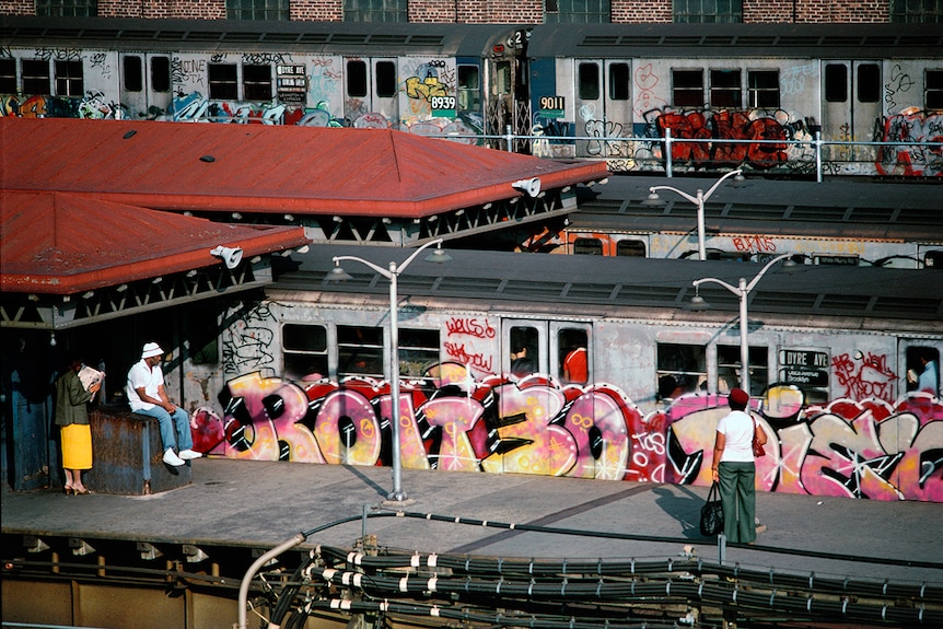 Three people wait for on subway station near three subway cars covered in colourful graffiti.