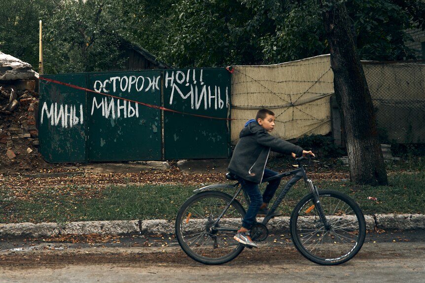Un adolescent fait du vélo sur une route, passant devant une clôture métallique avec des mots ukrainiens peints en blanc