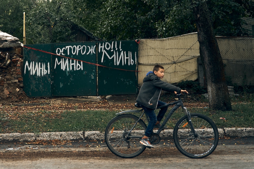 A teenager rides a bike down a road, passing a metal fence with ukrainian words painted on it in white