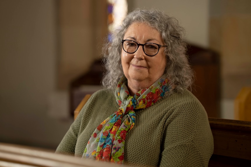 A woman wearing glasses sits in a pew inside a stone church, smiling wanly at the camera.