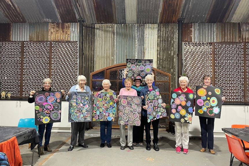 A group of ladies stand together holding paintings and smiling at the camera