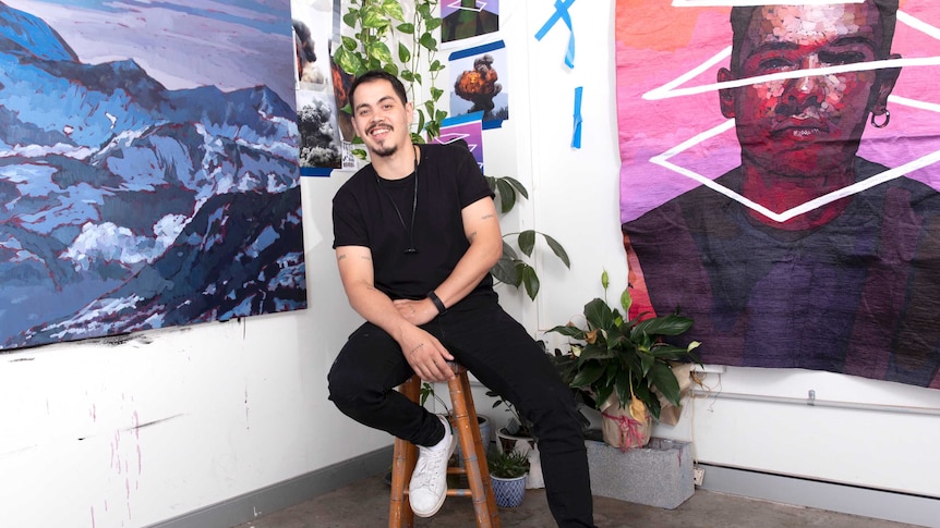 young man with facial hair smiling at camera in laidback pose between two large artworks