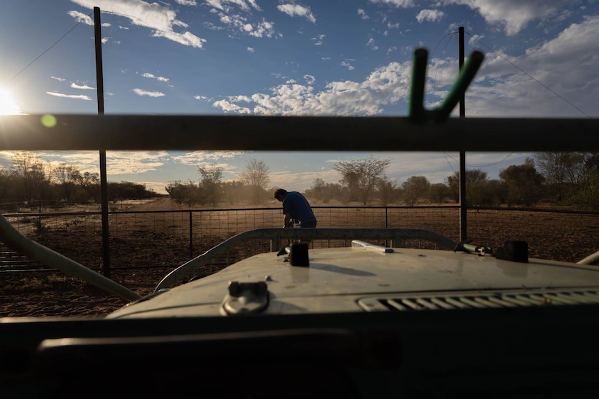 A shot from inside an old Jeep looking towards a gate on a property. A silhouette of a man leans over to open the gate.