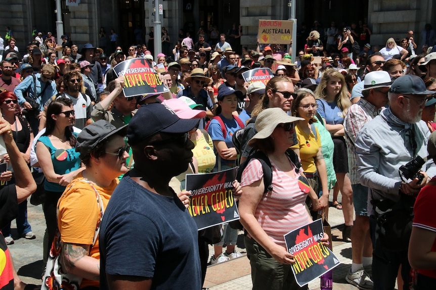 People in a large crowd hold signs and look towards a speaker as they protest.