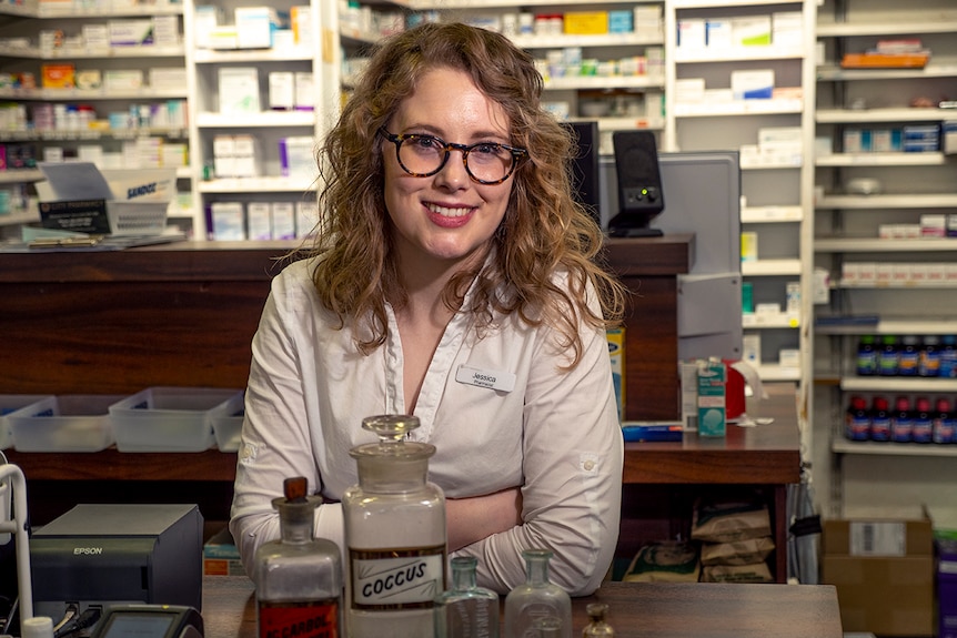 A young women stands at a pharmacy counter surrounded by medicines and some very old medicine bottles