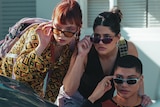 Three teens perch behind a car, bent over and spying on someone. They all are lowering their sunglasses for a closer look.