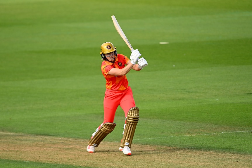 Ellyse Perry wearing a gold coloured helmet and pads and orange team colours hits down the ground during a Hundred T20 match.