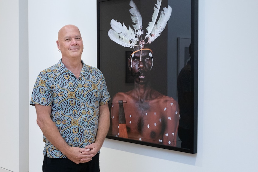 A man smiles as he stands next to a photo of an Indigenous man in ceremonial dress.