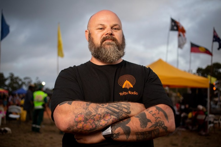 Man with bald head and crossed arms stands wearing black shirt with crowd and dark clouds visible in background.