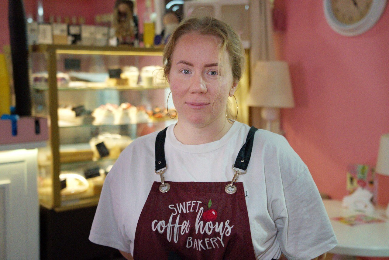 A woman wearing an aprin looks at the camera. A cabinet with cakes in it is visible behind her.