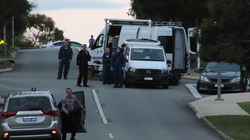 Forensic officers stand on the road next to a couple of large white vans. There is a plain-clothes officer in the foreground.