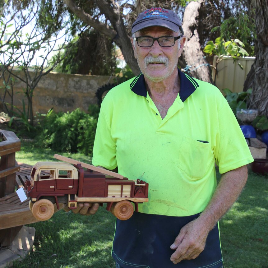 A man in a yellow hi-vis shirt and blue cap stands in his backyard holding a small wooden fire engine.