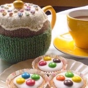 A bright yellow tea pot and tea cup on a white table cloth with three iced cupcakes on a plate.