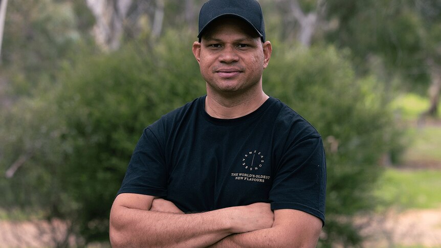 A man smiles for a photo wearing a hat and black shirt with his arms crossed. He is outside.