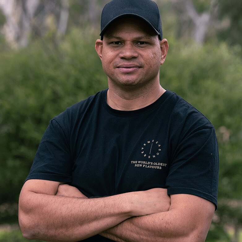 A man smiles for a photo wearing a hat and black shirt with his arms crossed. He is outside.