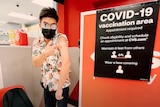 A man in a black face mask and sunglasses points to his shoulder next to a 'COVID-19 vaccination area' sign