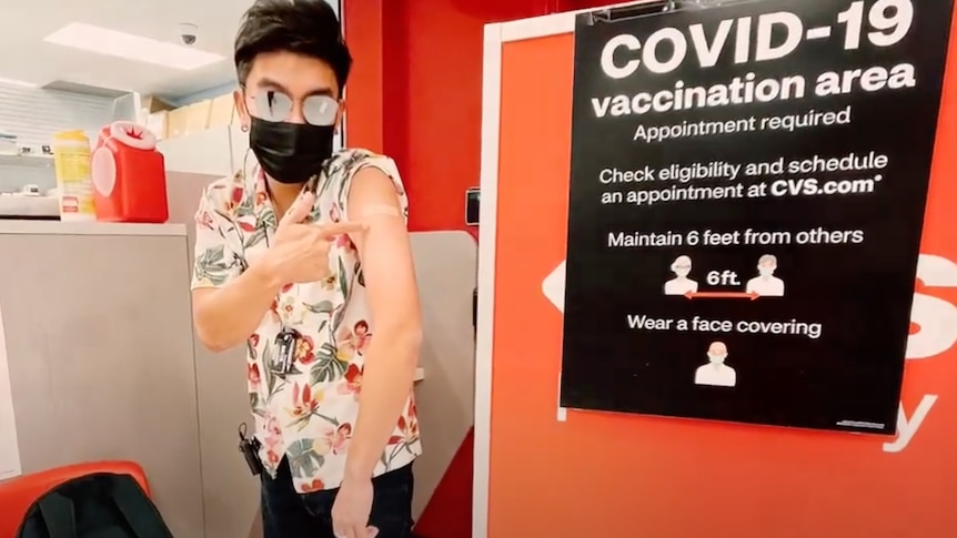 A man in a black face mask and sunglasses points to his shoulder next to a 'COVID-19 vaccination area' sign