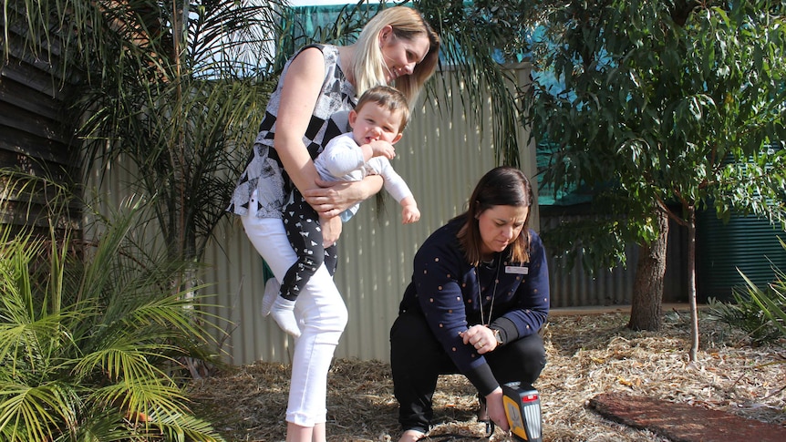 A mother smiles as she holds her baby while a health professional measures metals in the soil.