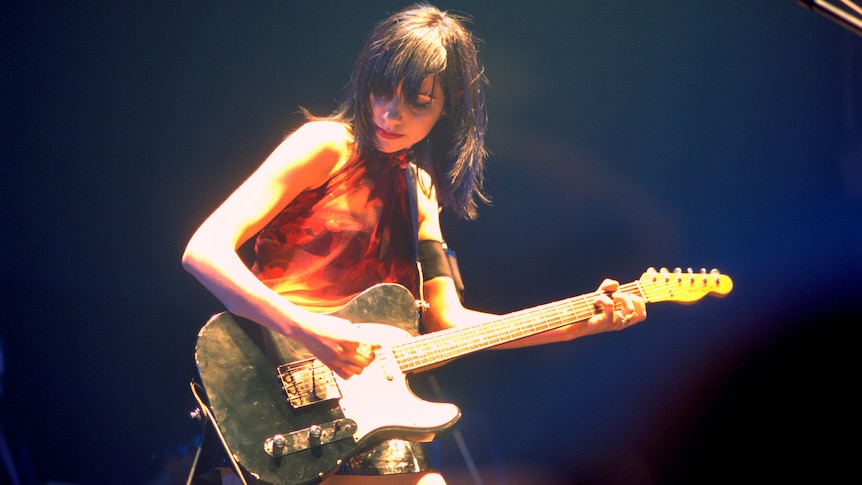 PJ Harvey, wearing a red dress, plays guitar on stage